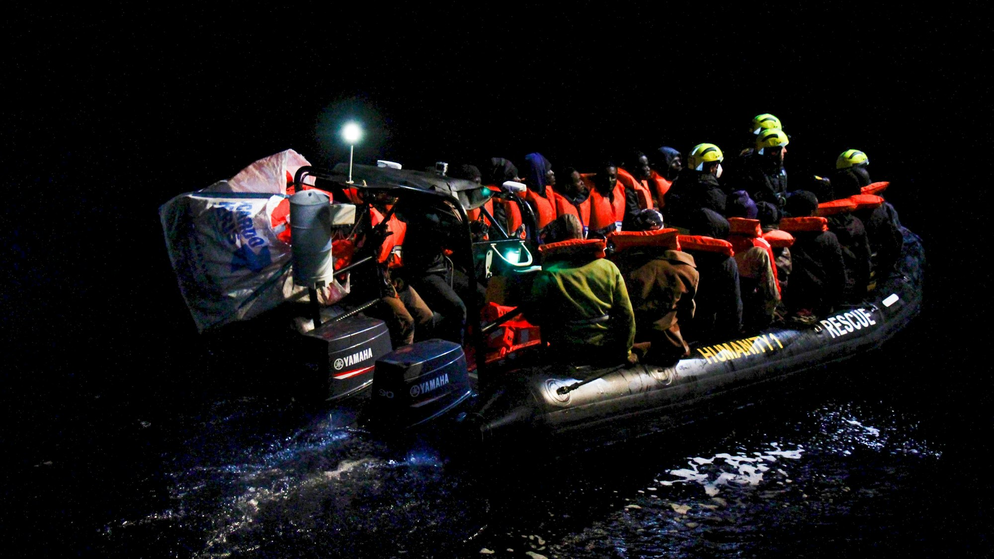 Menschen mit roten Rettungswesten sitzen in einem Schlauchboot auf dem Wasser. Es ist Nacht, Strahler sorgen für Licht.