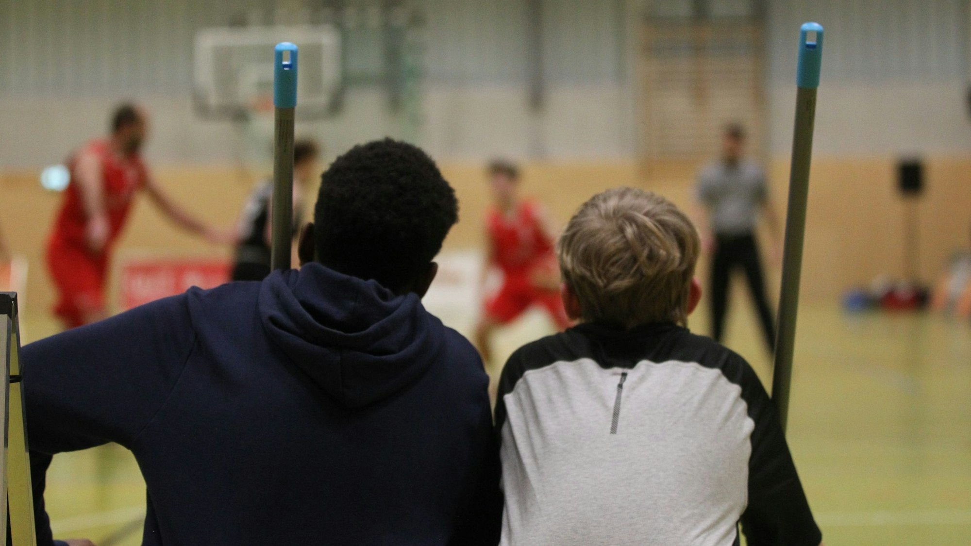 Die beiden Wischer werden von hinten gezeigt, die Stiele ihrer Besen überragen sie. Im Hintergrund sieht man verschwommen das Spielgeschehen.