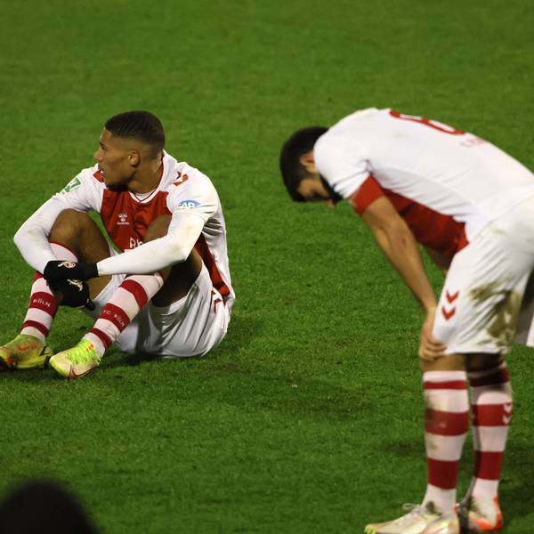 Ein Fußballspieler sitzt nach einer Niederlage enttäuscht auf dem Rasen, ein anderer stützt die Hände auf die Knie.
