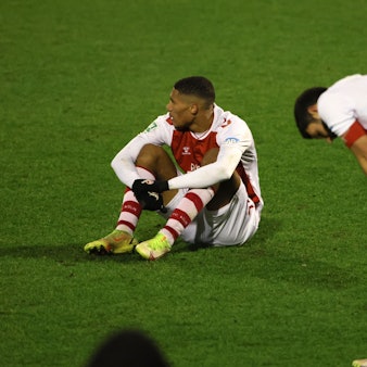 Ein Fußballspieler sitzt nach einer Niederlage enttäuscht auf dem Rasen, ein anderer stützt die Hände auf die Knie.