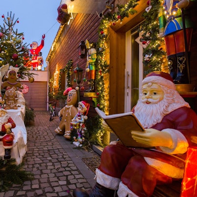 Mehrere Weihnachtsmänner, aber auch ein Engel und Lebkuchenfiguren stehen vor einem holzgetäfelten Haus. Auch ein geschmückter Weihnachtsbaum und ein Nikolaus auf einem Garagendach sind zu sehen.