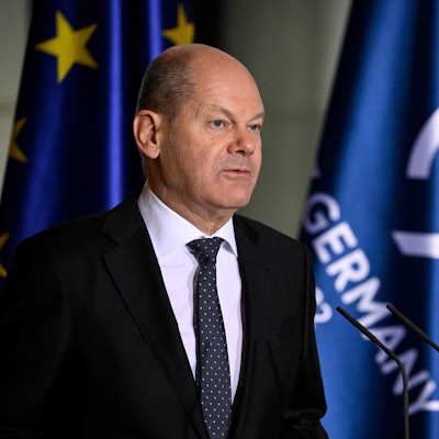 Olaf Scholz im Vordergrund steht vor Mikros und redet. Im Hintergrund sind die europäische Flagge und die der G7 zu sehen.