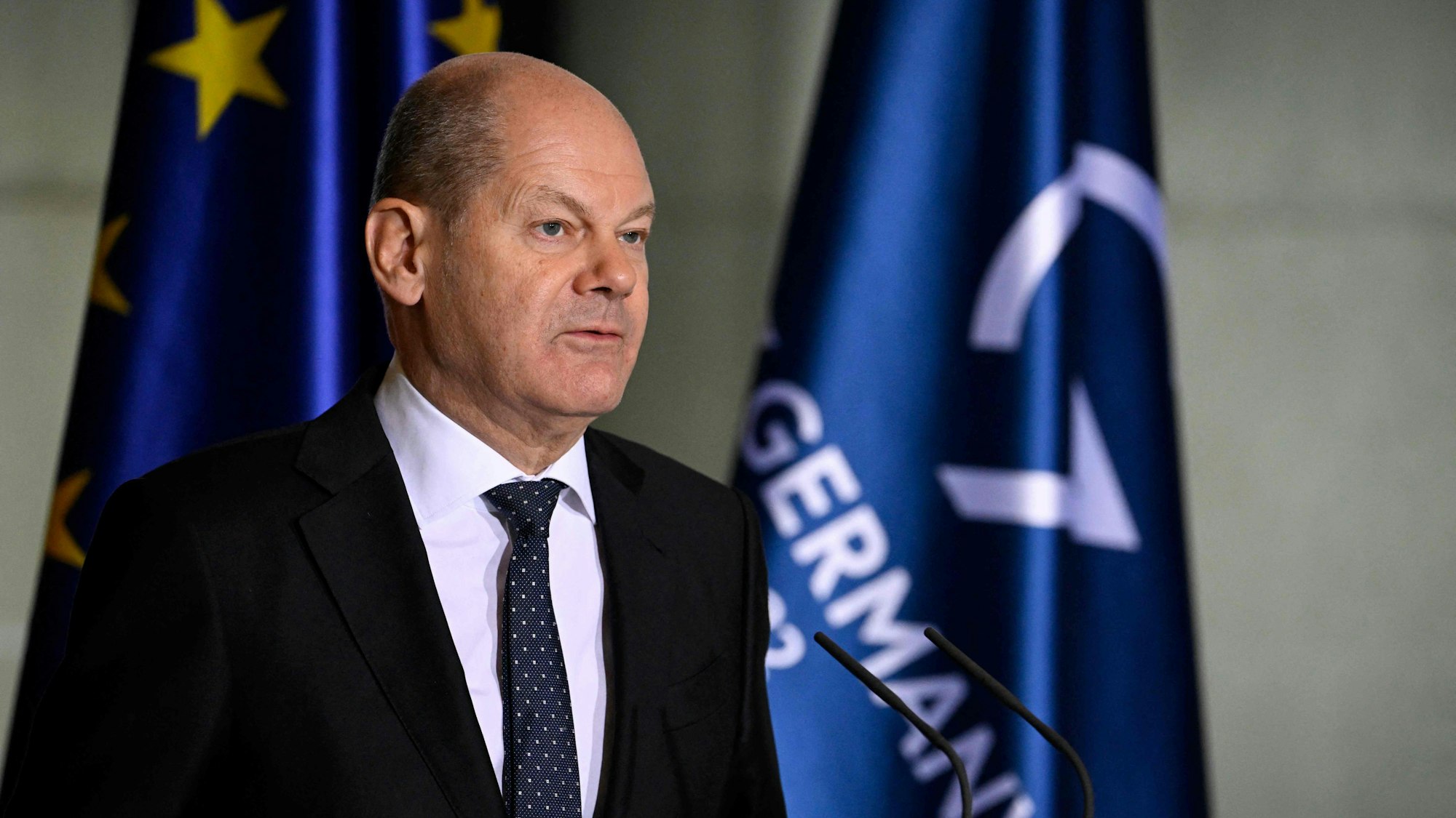 Olaf Scholz im Vordergrund steht vor Mikros und redet. Im Hintergrund sind die europäische Flagge und die der G7 zu sehen.