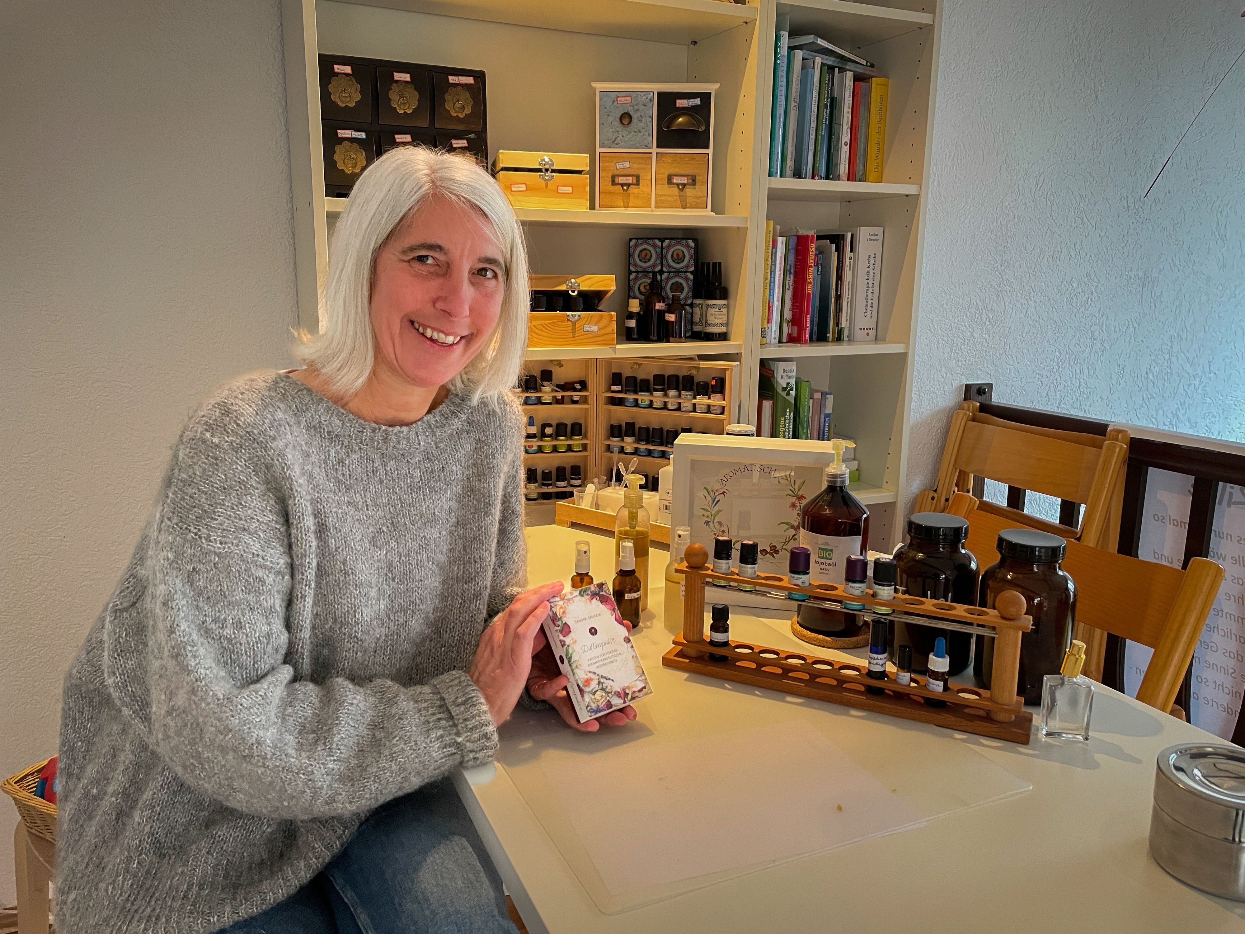 Aromaexpertin Doris Schultz hält ihr Buch in der Hand. Im Hintergrund stehen verschiedene Duftöle in einem Holzgestell.