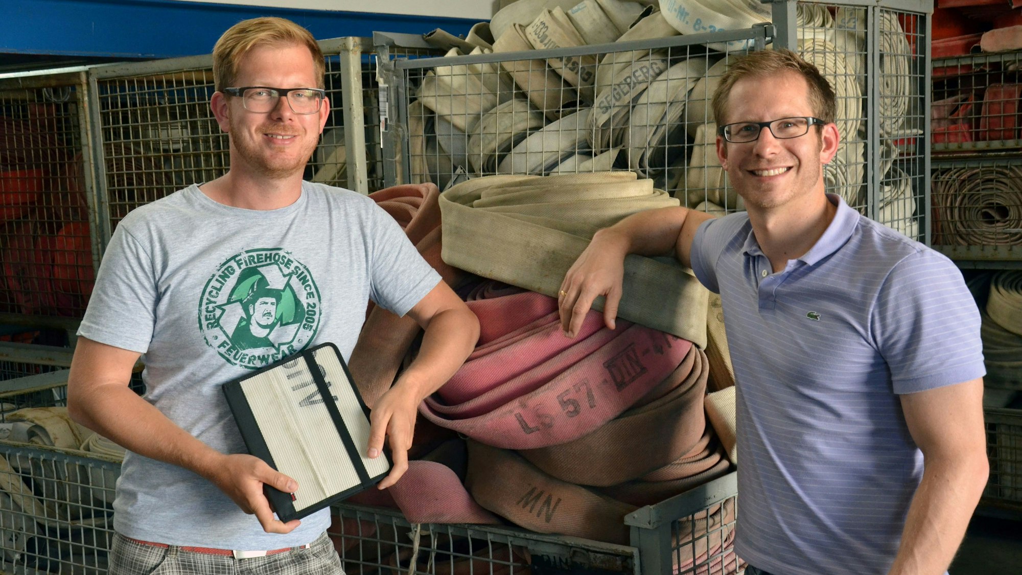 Martin und Robert Klüsener stehen neben Kisten voller alter Feuerwehrschläuchen und halten eine daraus hergestellte iPad-Hülle in der Hand.