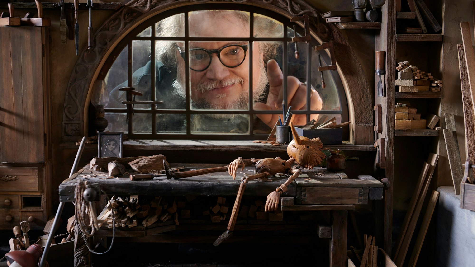 Regisseur Guillermo Del Toro steht hinter einer Fensterscheiber. Davor liegt Pinocchio aus seinem neuen Netflix-Film auf einer Werkbank.