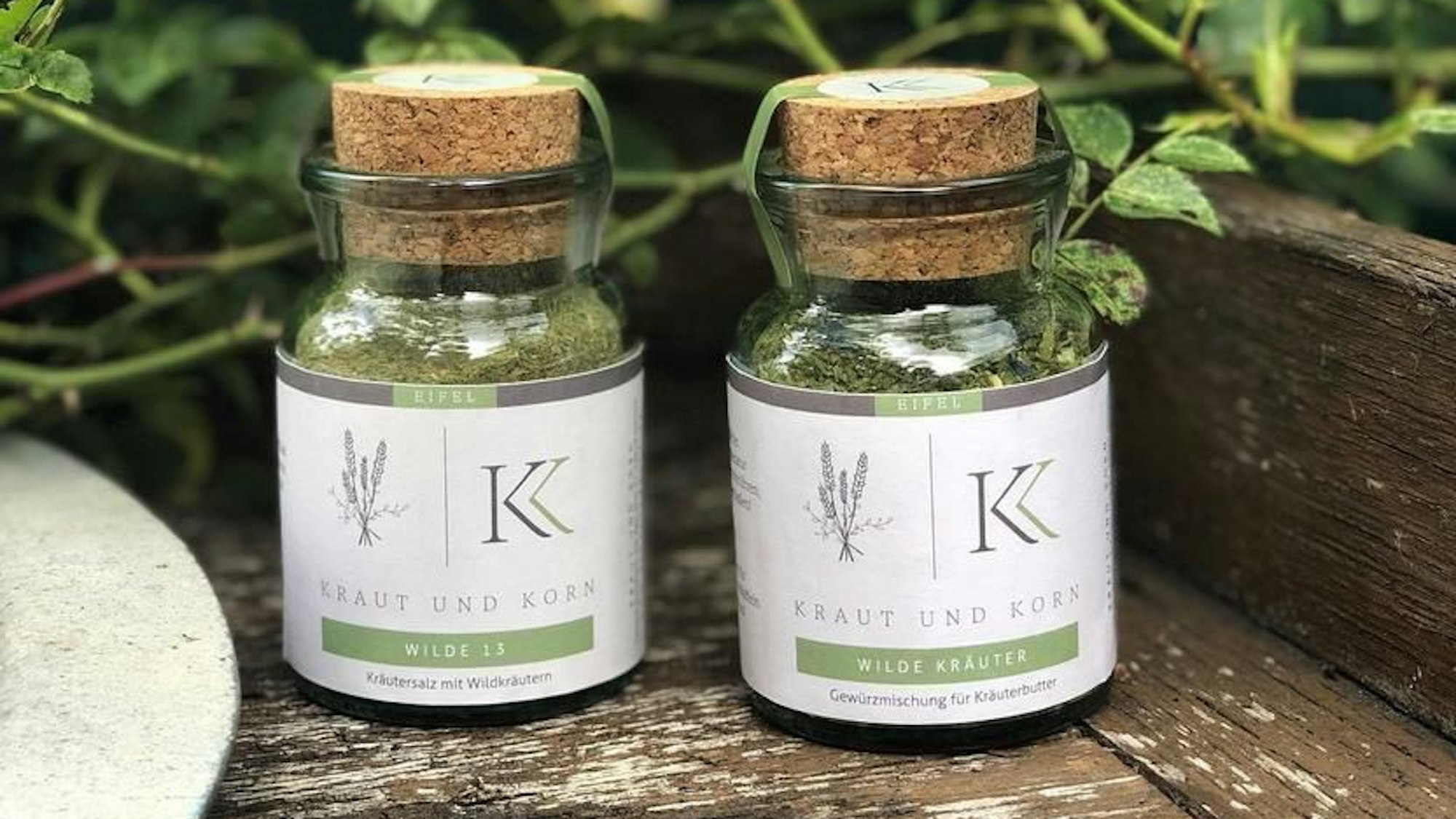 Zwei Gläser mit Gewürzmischungen aus der Manufaktur Kraut und Korn stehen auf einem Holzbrett, im Hintergrund sind grüne Pflanzen zu erkennen.