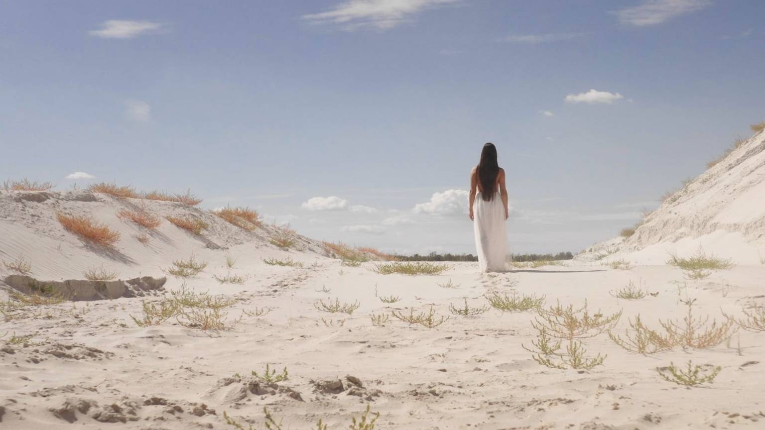 Eine junge Frau in einem langen weißen Kleid läuft durch eine Landschaft aus Sand.
