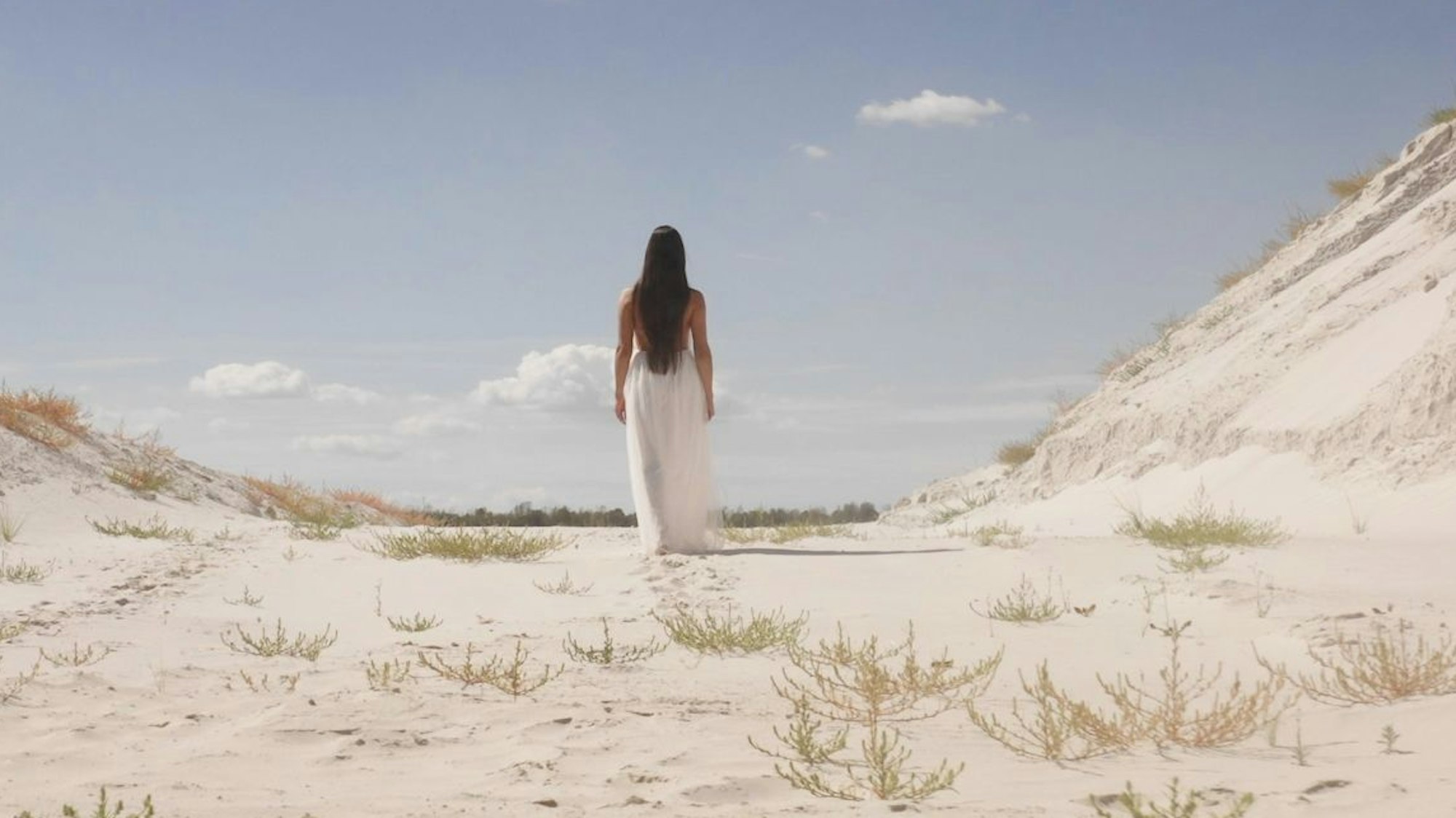 Eine junge Frau in einem langen weißen Kleid läuft durch eine Landschaft aus Sand.