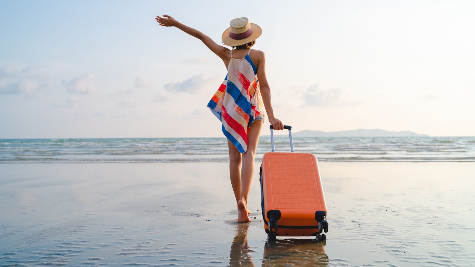 Eine Frau steht mit Rollkoffer am Strand.