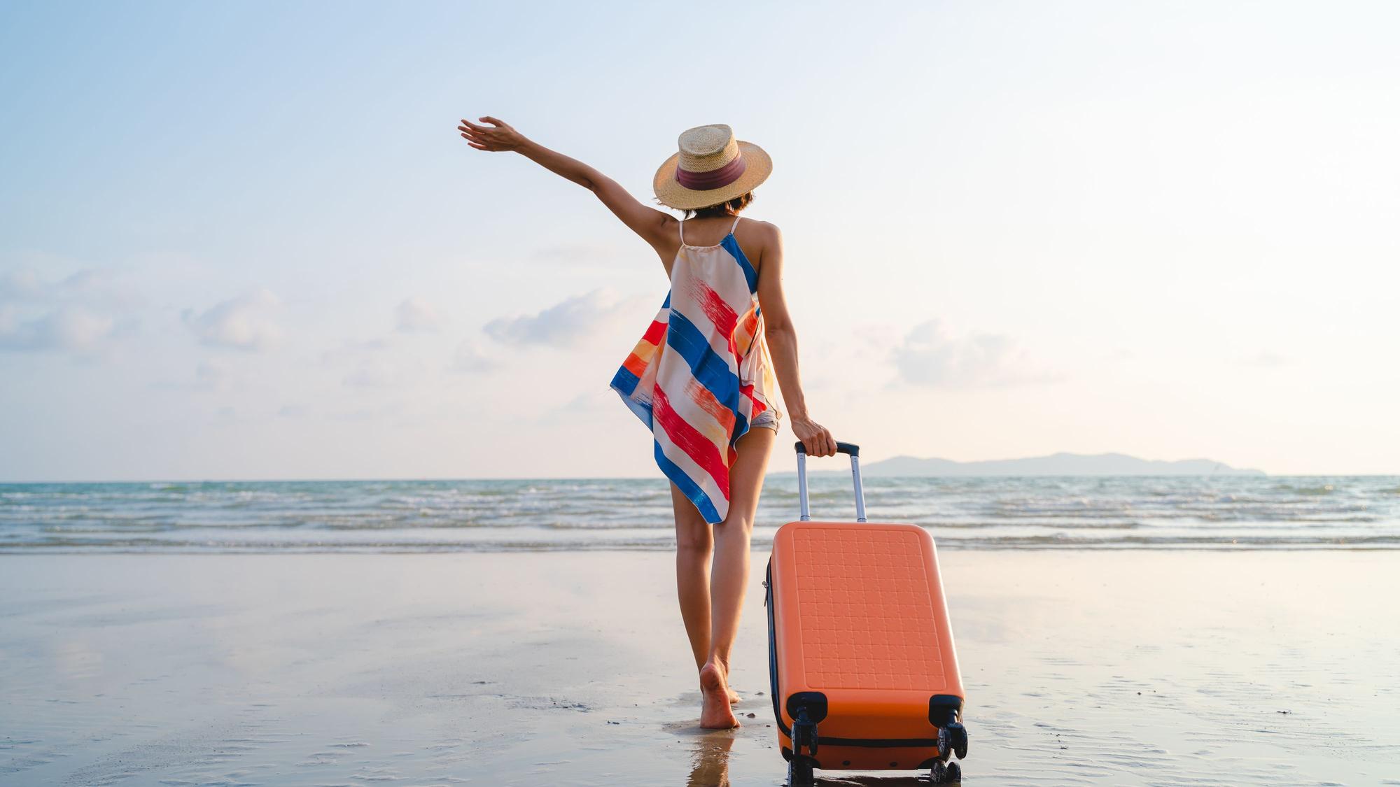 Eine Frau steht mit Rollkoffer am Strand.