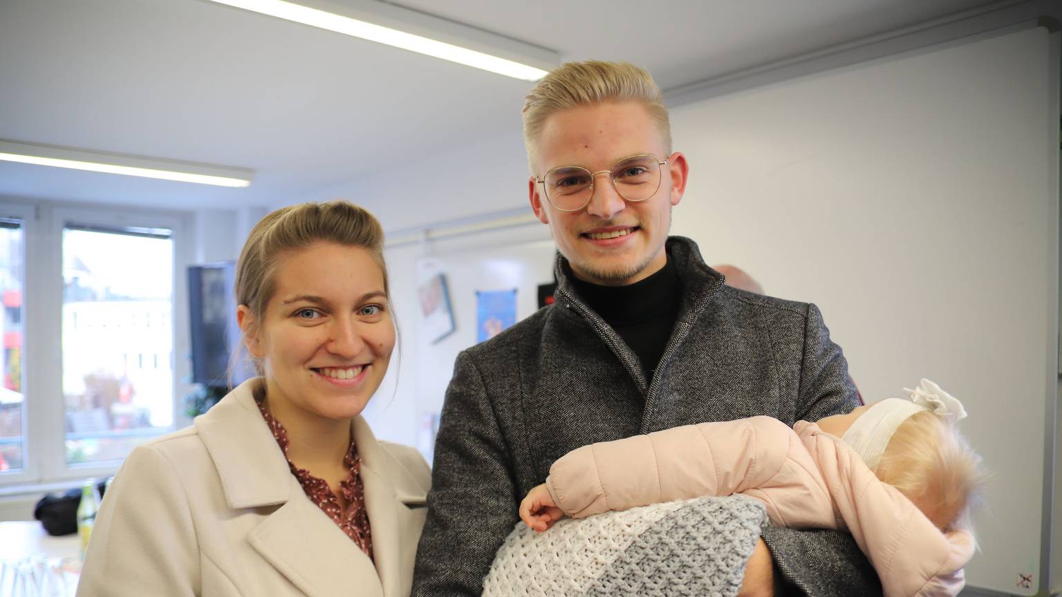 Eine Frau und ein Mann lachen in die Kamera. Der Mann hat ein Baby auf dem Arm.