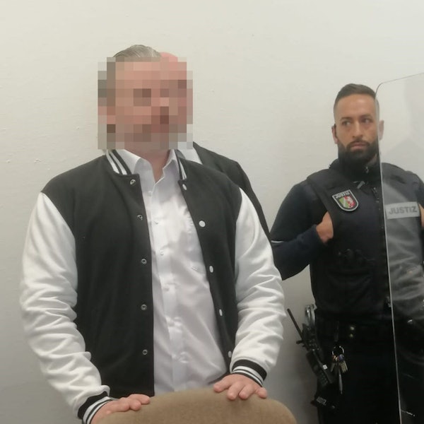 Das Bild zeigt den Angeklagten an der Seite eines Wachmanns im Gerichtssaal.