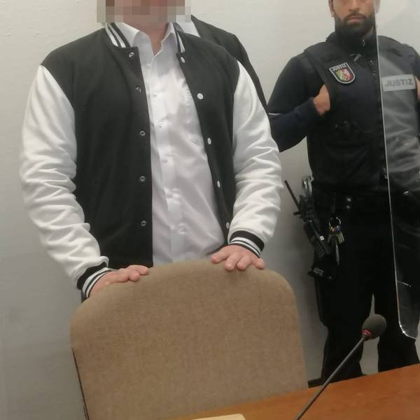 Das Bild zeigt den Angeklagten, der von einem Justizbeamten in den Gerichtssaal gebracht wird.
