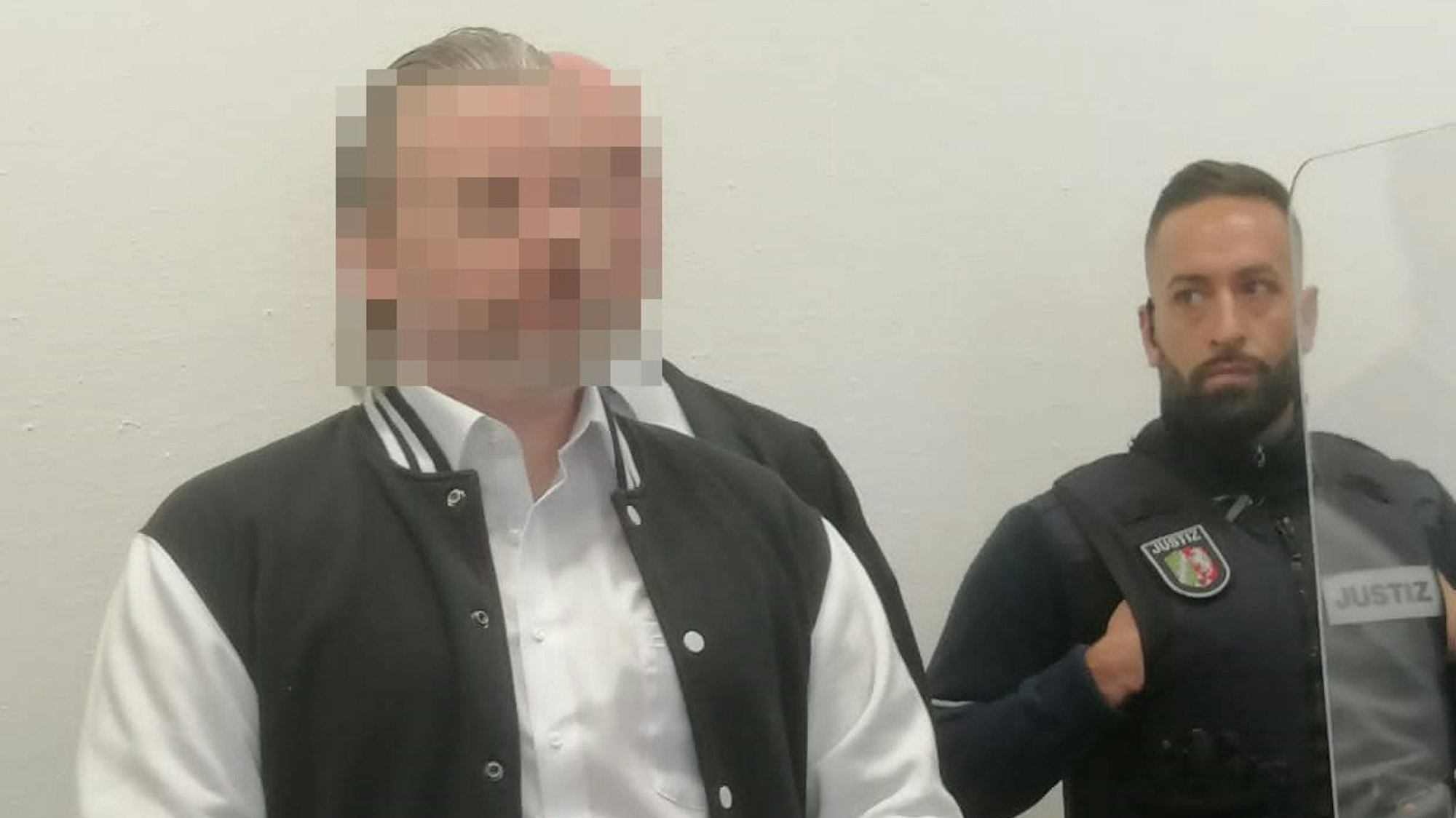 Das Foto zeigt das unkenntlich gemachte Gesicht des Angeklagten. Neben ihn steht ein Justizvollzugsbeamter.