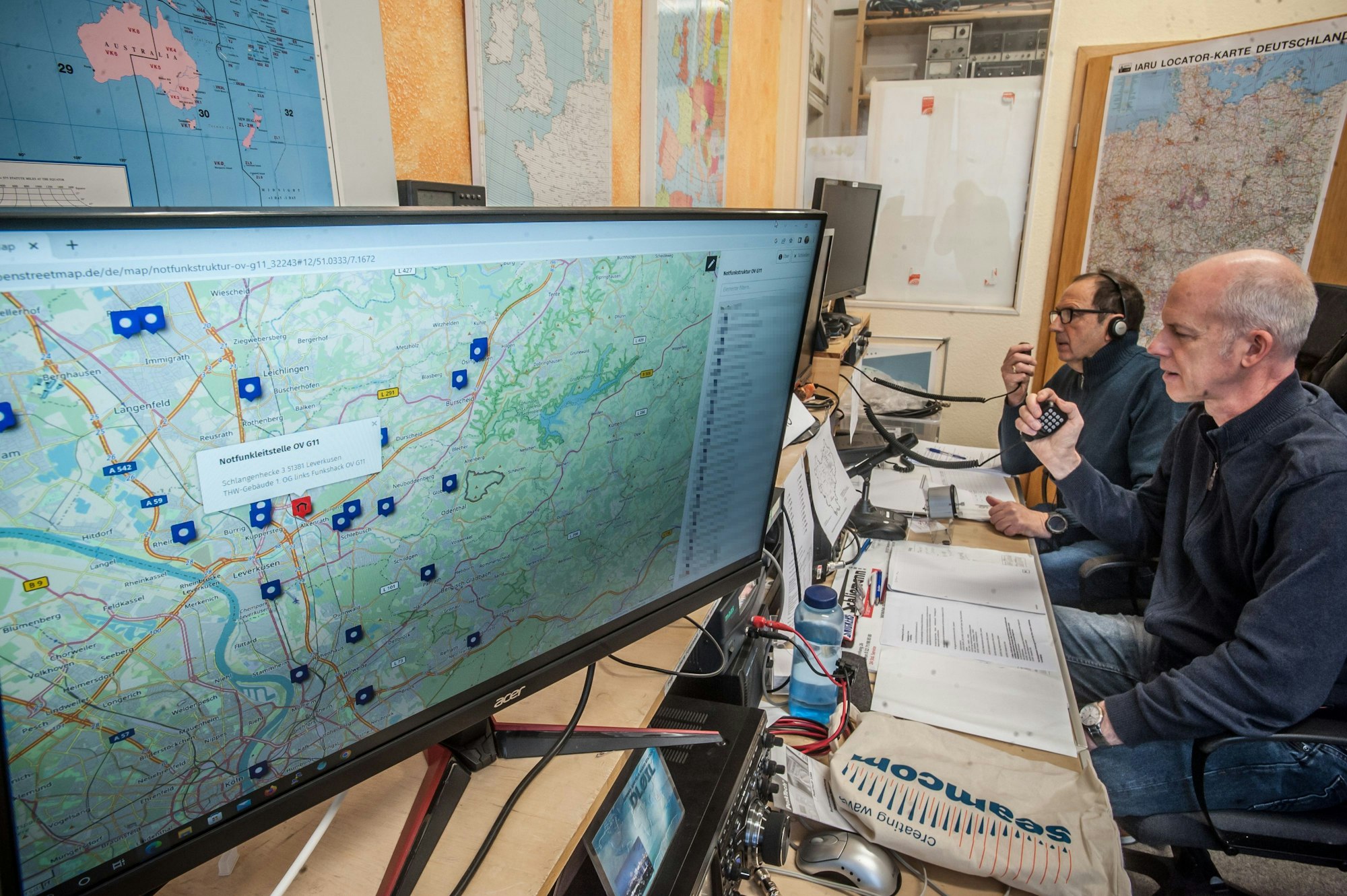 Simon Sauerwald und Achim Wagner sitzen vor Bildschirmen. In ihren Händen halten sie Funkgeräte.