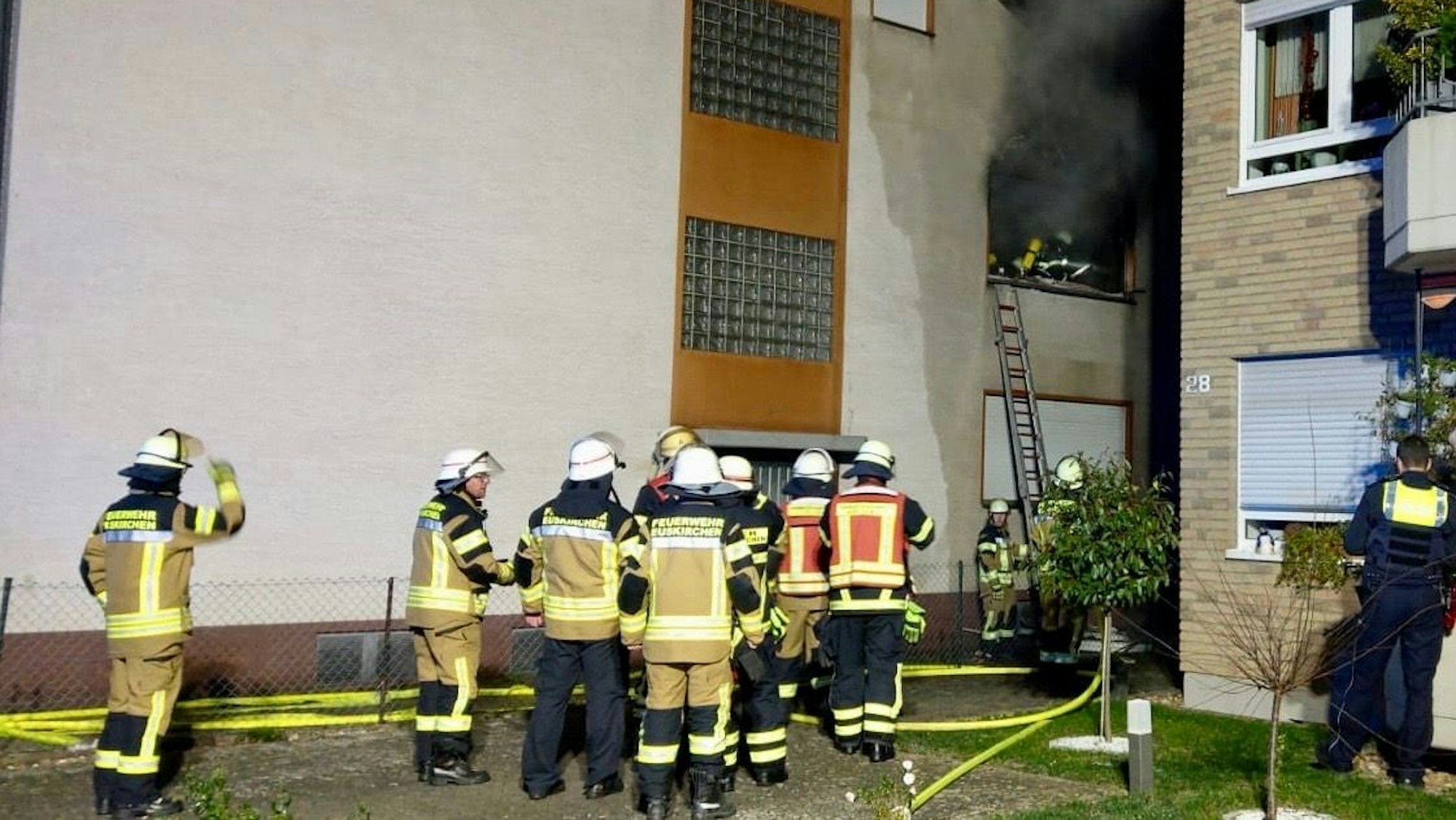 Feuerwehrleute stehen vor einem Mehrfamilienhaus. Eine Leiter ist an die Wand gelehnt. Aus dem Fenster kommt schwarzer Rauch.
