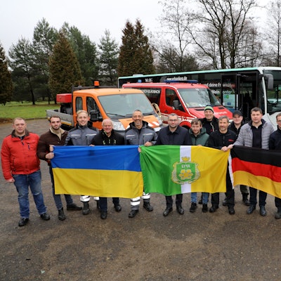 Eine Menschengruppe steht vor Bussen und hält drei Flaggen hoch.