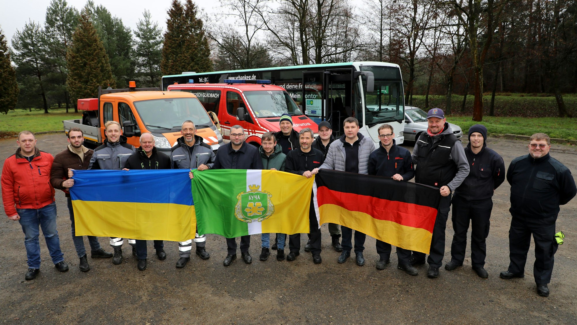 Eine Menschengruppe steht vor Bussen und hält drei Flaggen hoch.