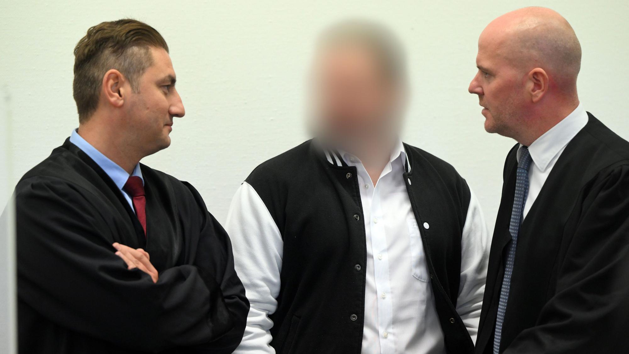 Der Angeklagte steht zwischen seinen Anwälten Mutlu Günal und Martin Bücher. Er trägt ein weißes Hemd und eine schwarz-weiße Sportjacke.