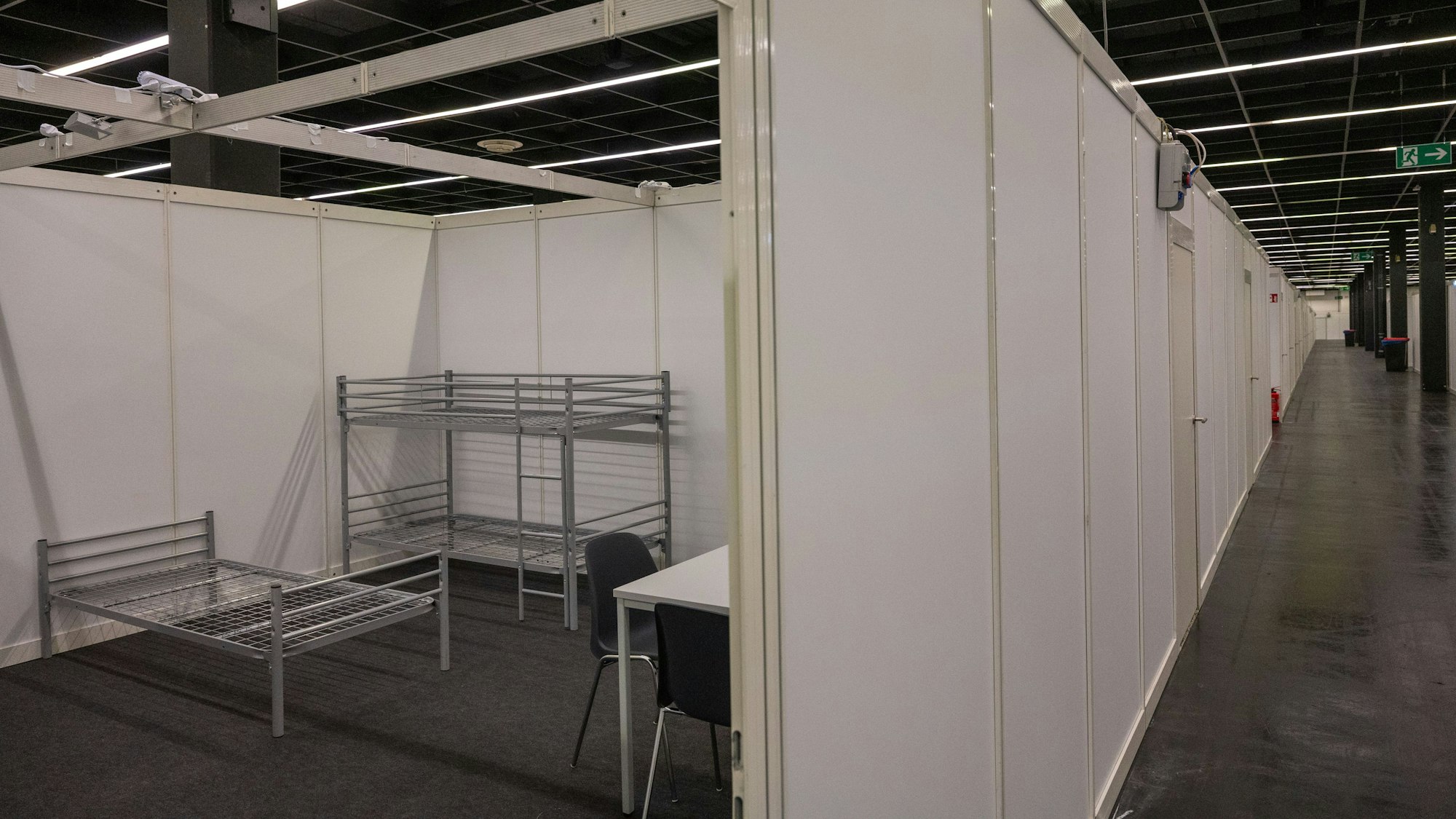 Eine Unterkunft für Geflüchtete in der Koelnmesse. Ein Bett und ein Hochbett sowie ein Tisch mit Stühlen steht auf der Fläche.