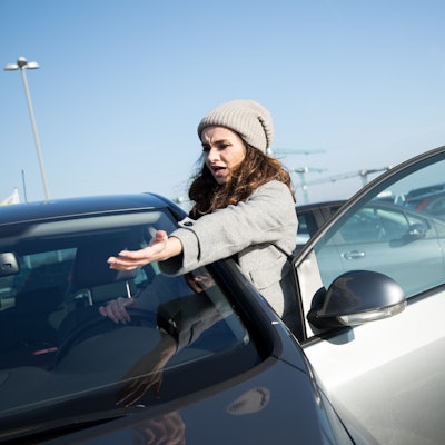 Eine Frau mit Mütze steht an ihrer Autotür und streckt wütend den Arm aus.
