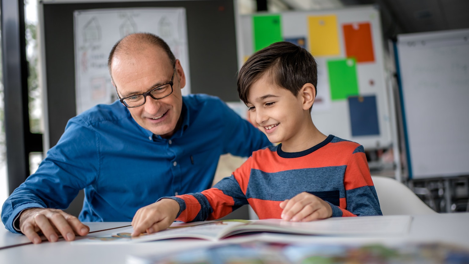 Mentor Andreas Endermann begleitet ein Kind beim Lesenlernen - das Bild zeigt die beiden, wie sie in einem Buch lesen, das vor ihnen auf dem Tisch liegt.