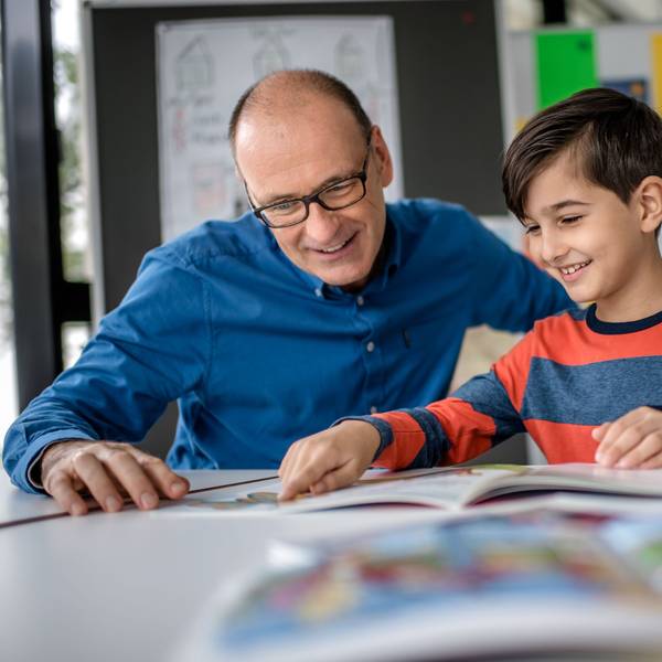 Mentor Andreas Endermann begleitet ein Kind beim Lesenlernen - das Bild zeigt die beiden, wie sie in einem Buch lesen, das vor ihnen auf dem Tisch liegt.