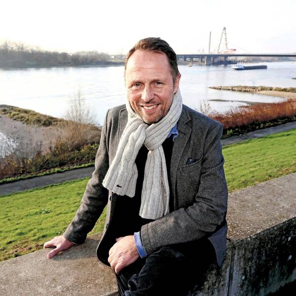 Uwe Richrath sitzt auf einer Mauer vor einer Böschung, die zum Rhein führt. Der Rhein ist groß im Hintergrund, ein Schiff ist zu sehen und die Leverkusener Rheinbrücke.