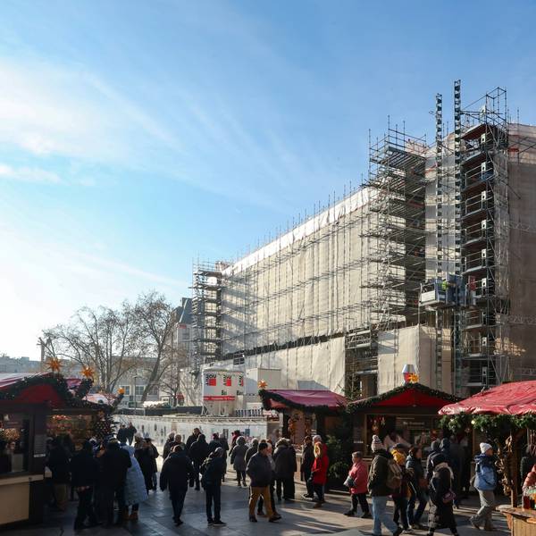 Das Bild zeigt das eingerüstete Dom-Hotel, davor stehen Buden des Weihnachtsmarktes auf dem Roncalliplatz.