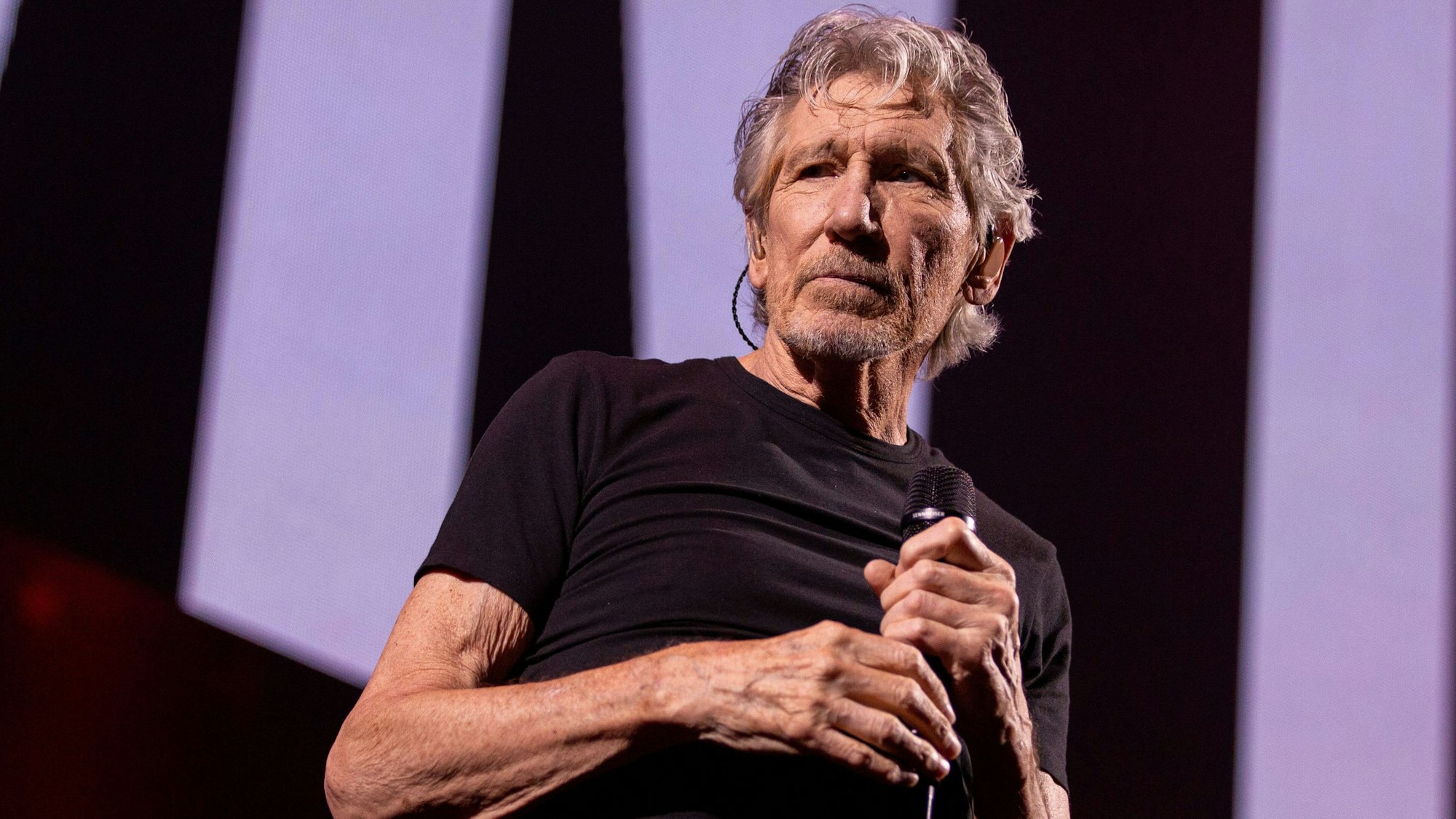 Roger Waters steht im schwarzen T-Shirt auf einer Konzertbühne.