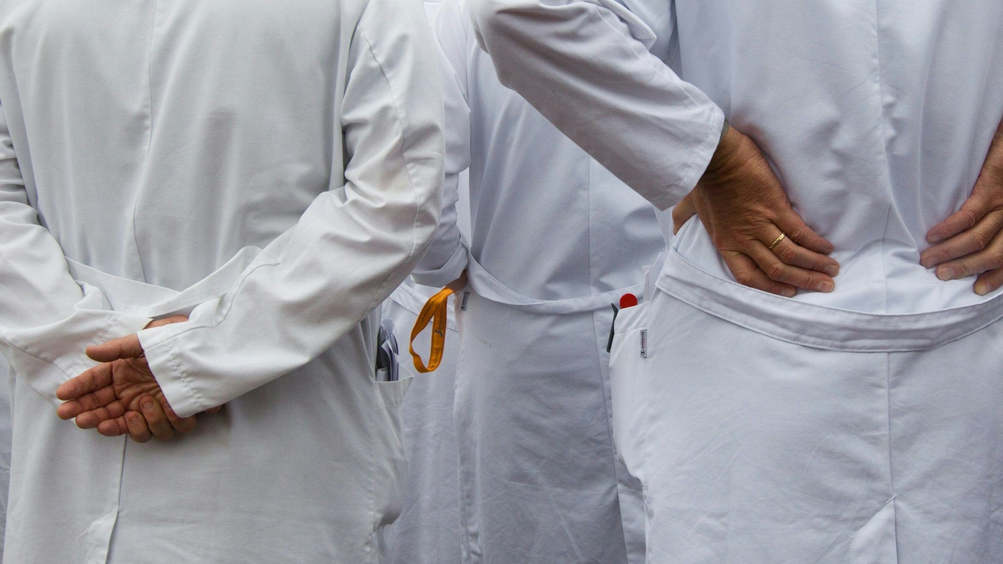Man sieht zwei Personen, die der Kamera den Rücken zugedreht haben. Sie tragen weiße Kittel, die eine Person hat die Hände in die Hüften gestemmt, die andere verschränkt sie auf dem Rücken. Links uns rechts sind andere Personen teilweise zu sehen.