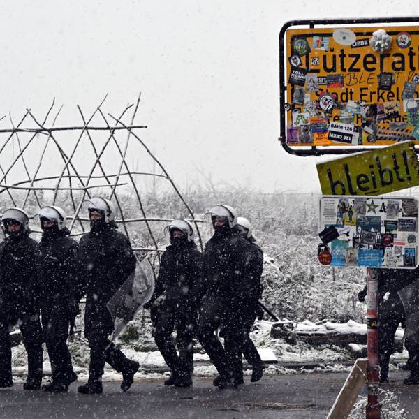 Polizisten stehen bei dem mit Stickern zugeklebten Ortsschild zum Eingang von Lützerath. Um sie herum liegt Schnee.