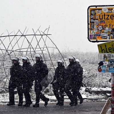 Polizisten stehen bei dem mit Stickern zugeklebten Ortsschild zum Eingang von Lützerath. Um sie herum liegt Schnee.