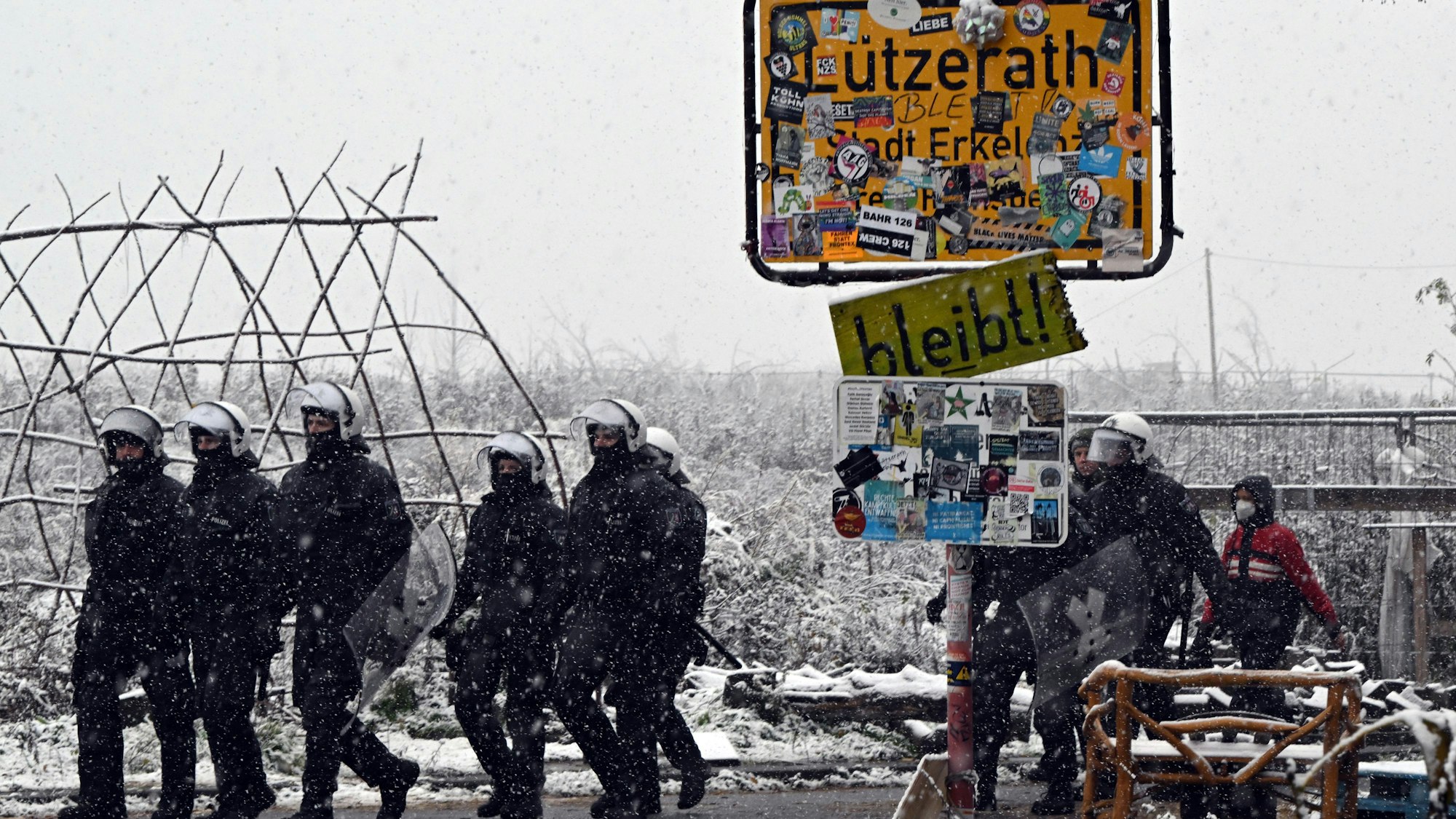 Polizisten stehen bei dem mit Stickern zugeklebten Ortsschild zum Eingang von Lützerath. Um sie herum liegt Schnee.