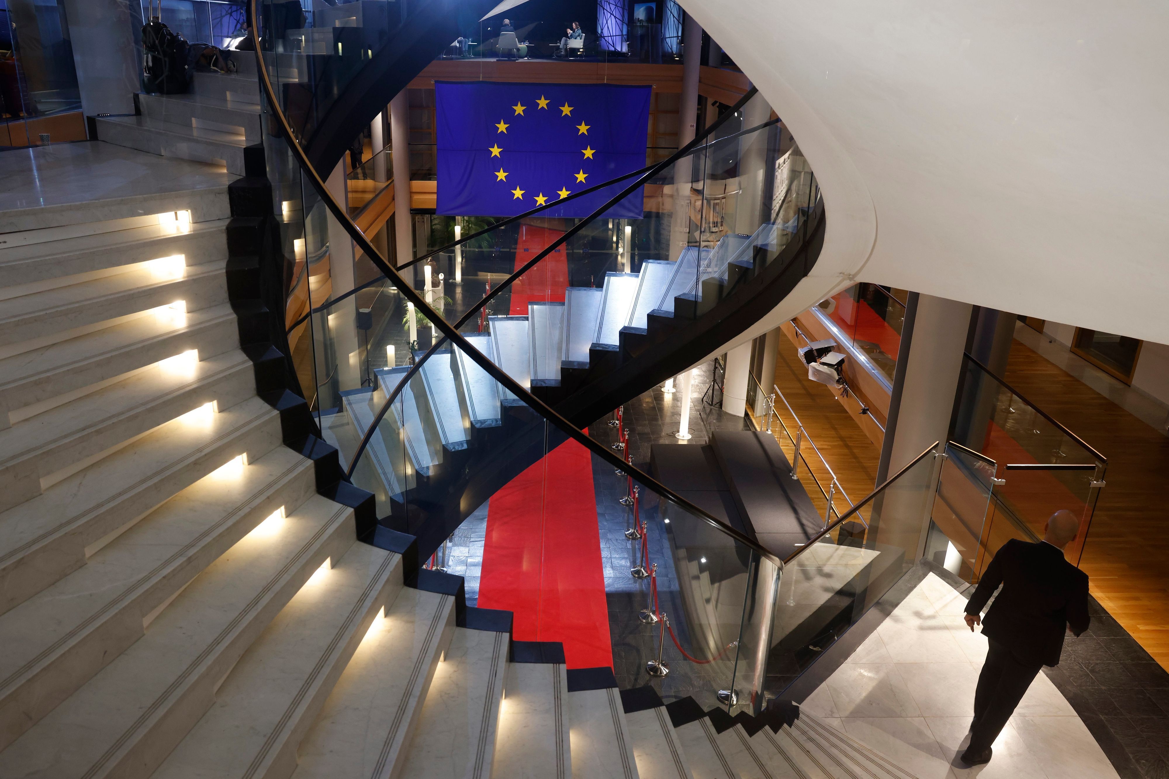 Zu sehen ist ein Treppenhaus mit zwei  beleuchteten Wendeltreppen. Der Blick geht nach unten in eine Halle, in der ein roter Teppich liegt, darüber hängt die Flagge der EU.