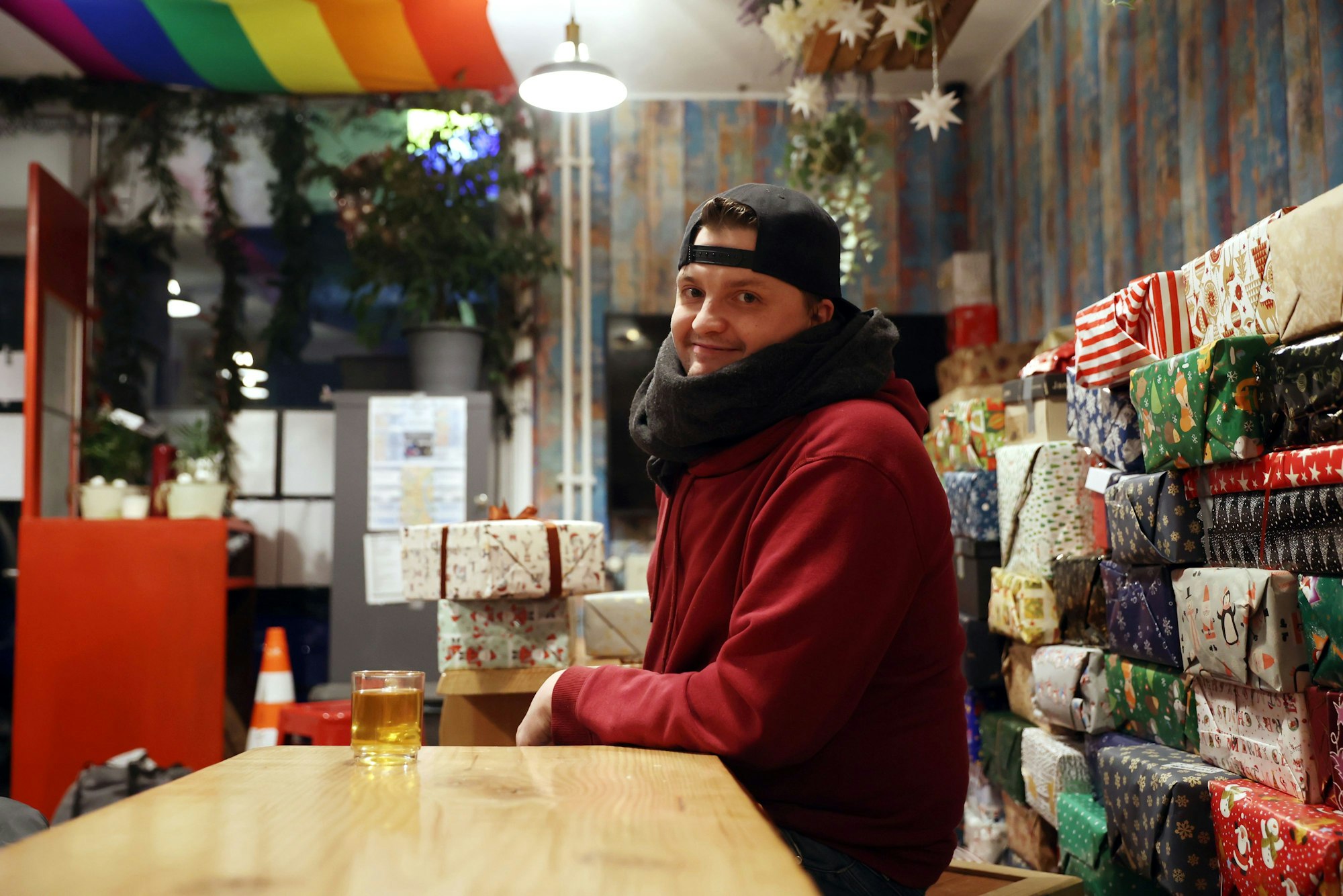 Ein junger Mann sitzt in rotem Pulli und dickem Schal an einem Tisch vor aufgestapelten Geschenken.