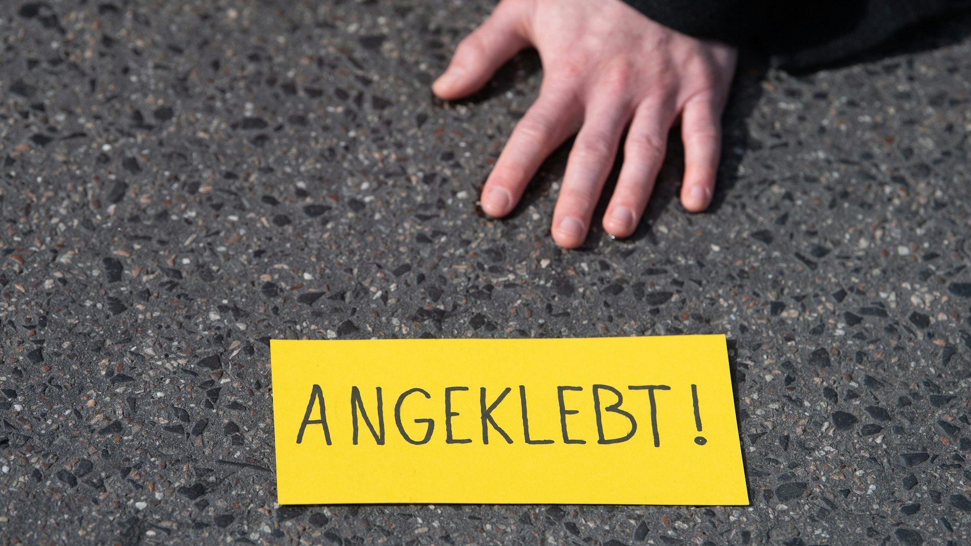 Zu sehen ist der Ausschnitt einer Betonoberfläche. Auf ihr liegt eine Hand, darunter liegt ein gelber Zettel mit der Aufschrift „Angeklebt“.