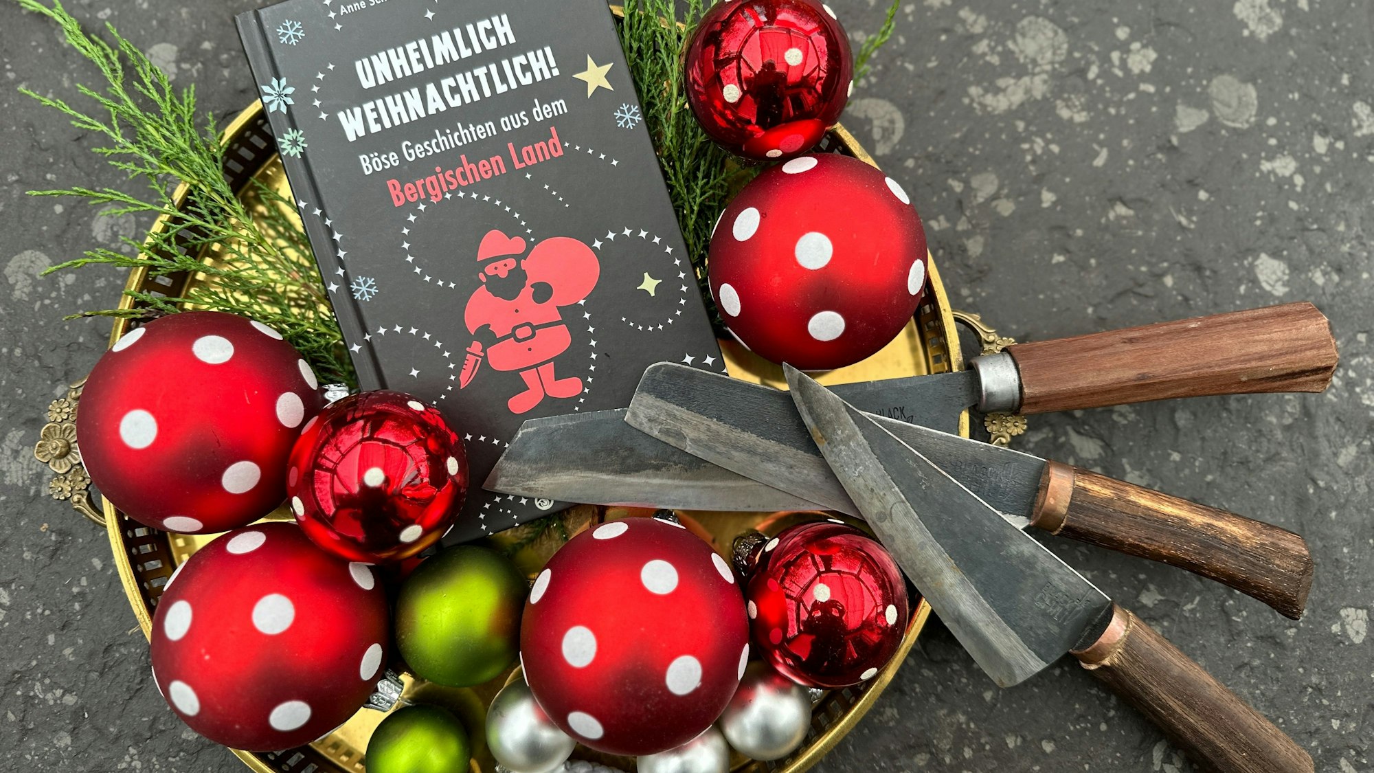 Das Buch „Unheimlich weihnachtlich – Böse Geschichten aus dem Bergischen Land“ liegt in einer Schale mit Weihnachtskugeln - und drei Messern.