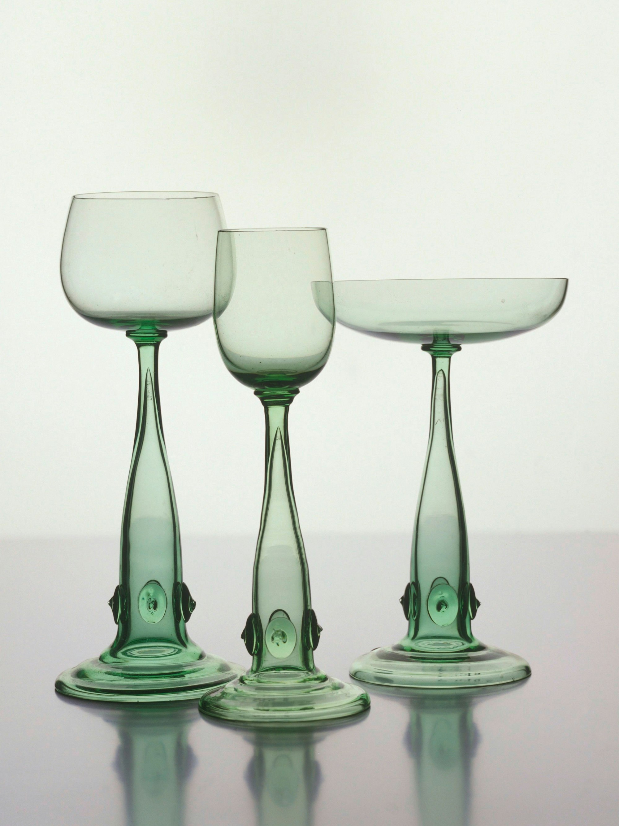 Drei grünlich schimmernde Tafelgläser mit Trompetenfüßen stehen vor einer weißen Wand.