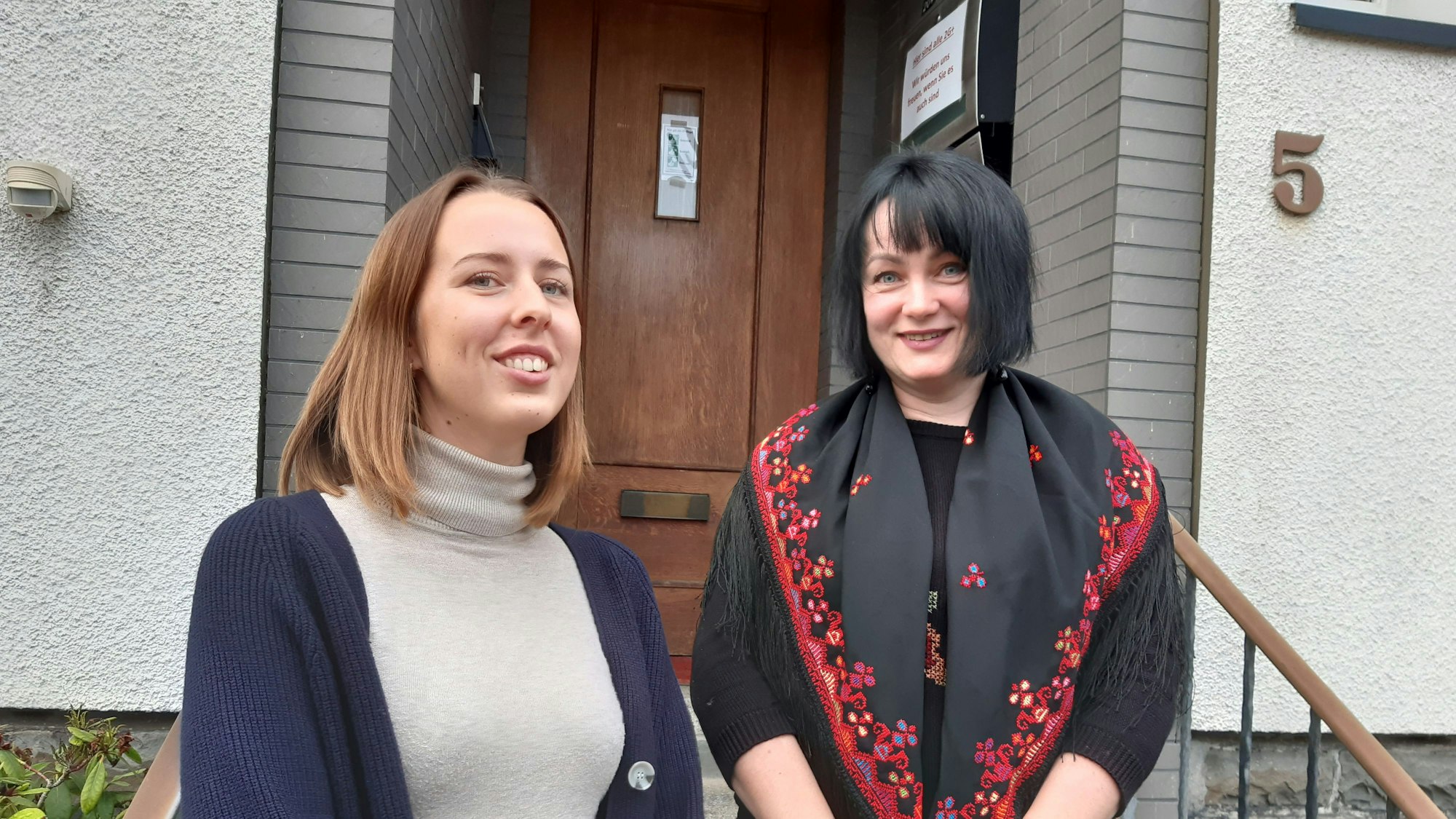 Zwei Frauen stehen vor einer Tür mit der Hausnummer fünf und lächeln in die Kamera.