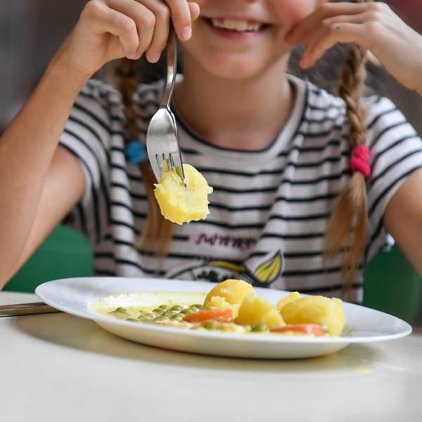 Eine Schülerin sitzt in einer Schulmensa vor einem Teller mit Kartoffeln, Erbsen und Möhren.