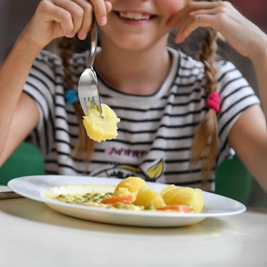 Eine Schülerin sitzt in einer Schulmensa vor einem Teller mit Kartoffeln, Erbsen und Möhren.