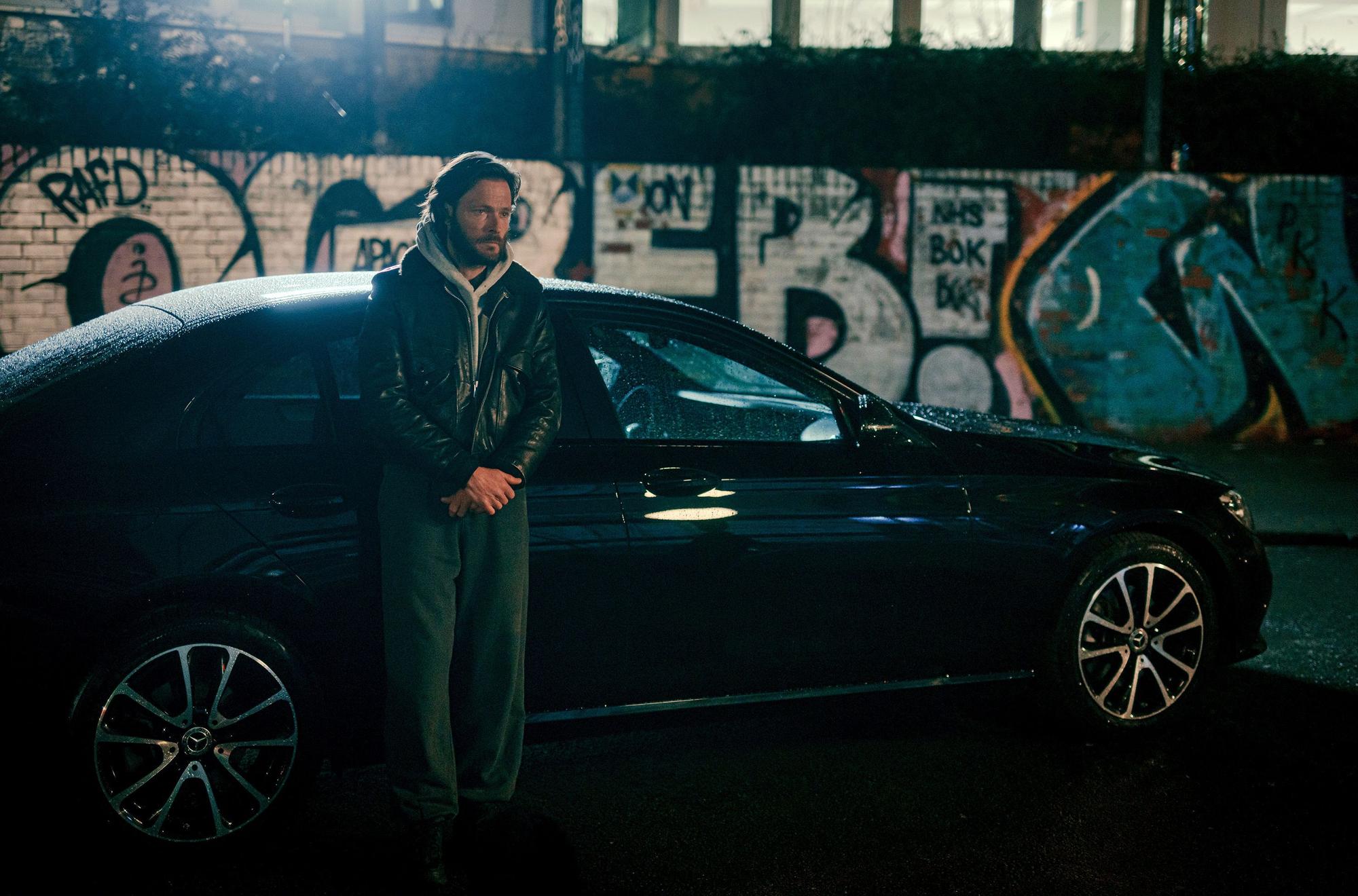 Maik steht vor seinem schwarzen Auto und wartet auf Sammy. Im Hintergrund ist eine mit Graffiti besprühte Wand.