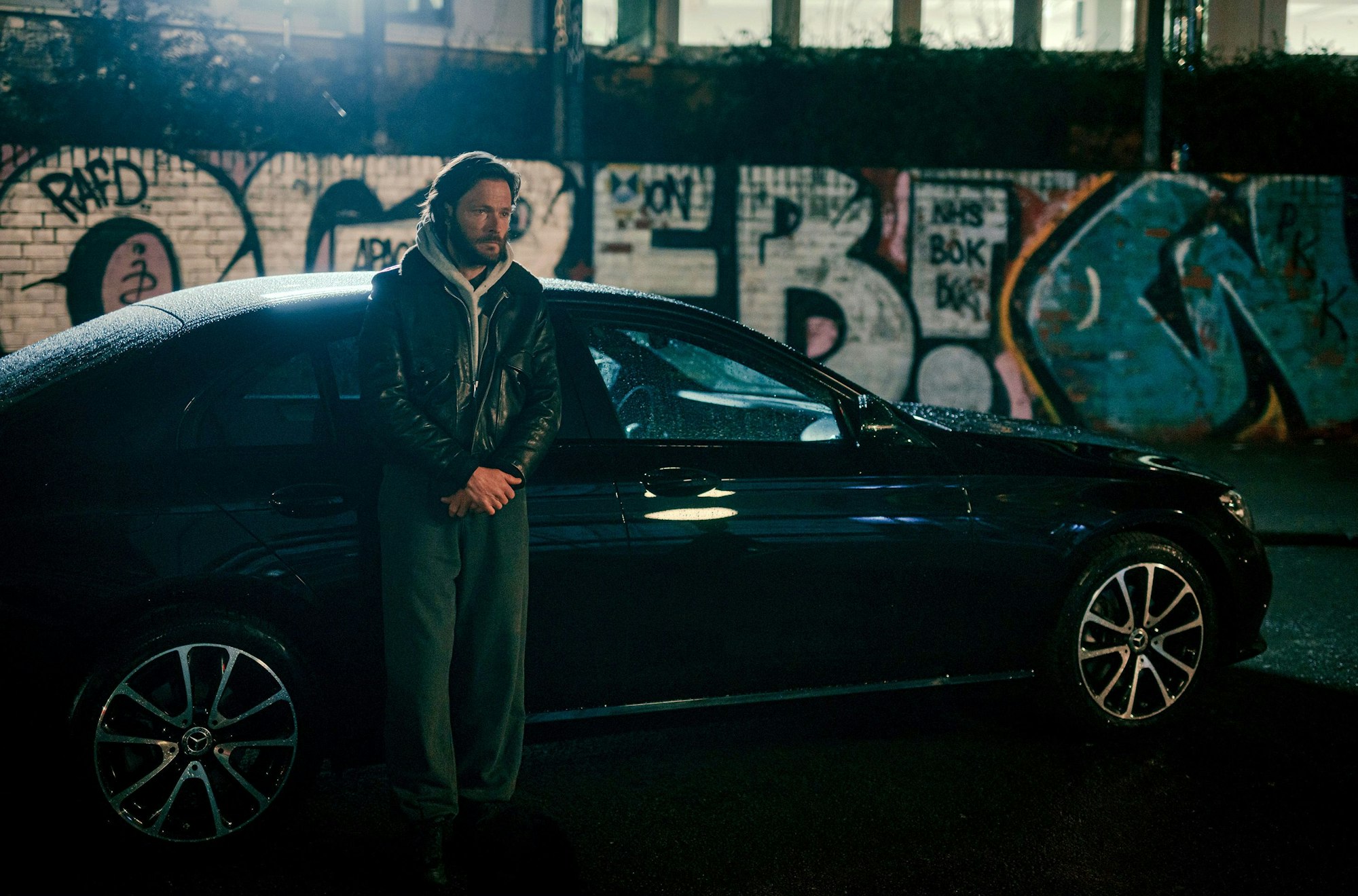 Maik steht vor seinem schwarzen Auto und wartet auf Sammy. Im Hintergrund ist eine mit Graffiti besprühte Wand.