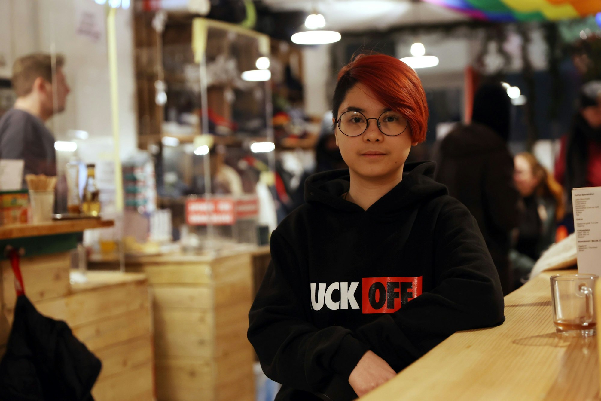 Dino steht in schwarzem Sweatshirt und rot gefärbten Haaren an einer Holztheke.