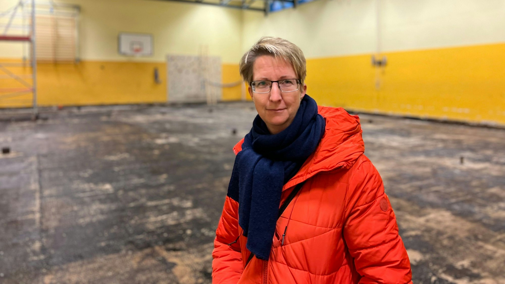 Jana Herrmann, Vertreterin der Schulpflegschaft der KGS Kupfergasse in Urbach, steht in der maroden Turnhalle der Schule. Der Bode ist schwarz, hinter ihr ist ein Gerüst für die Arbeiten aufgebaut.