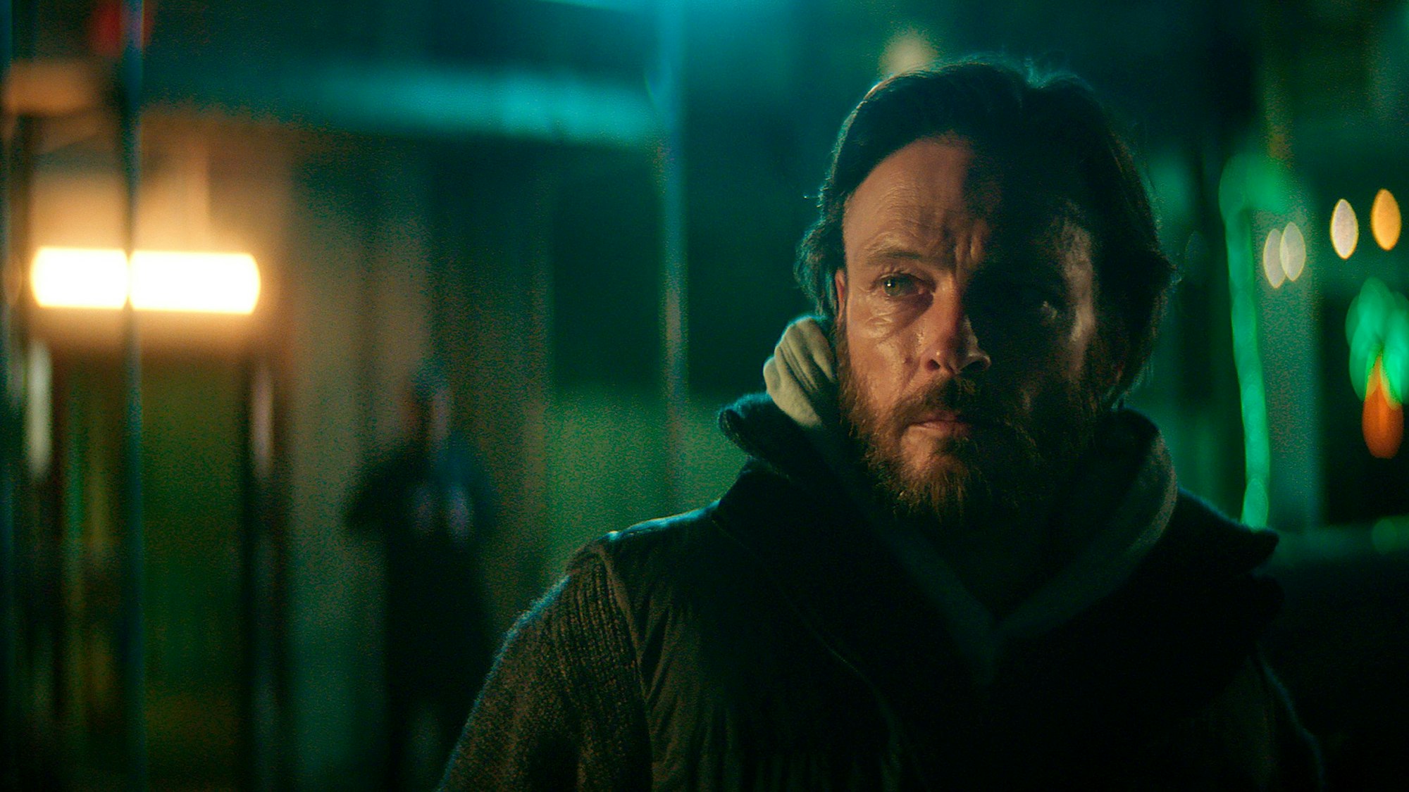 Andreas Pietschmann als Maik Balthasar, er trägt einen Kapuzzenpulli und darüber eine Jacke. Der Hintergrund, das Berliner Nachtleben, ist unfokussiert.