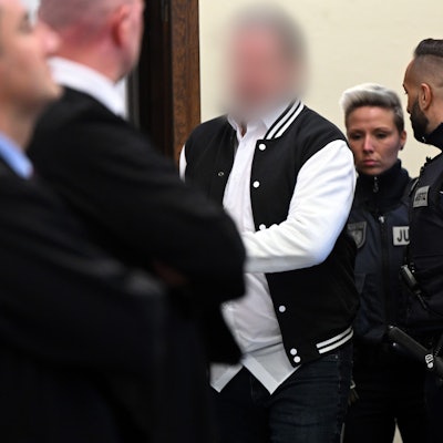 Der Angeklagte (m) wird von mehreren Justizbeamten in den Gerichtssaal geführt.