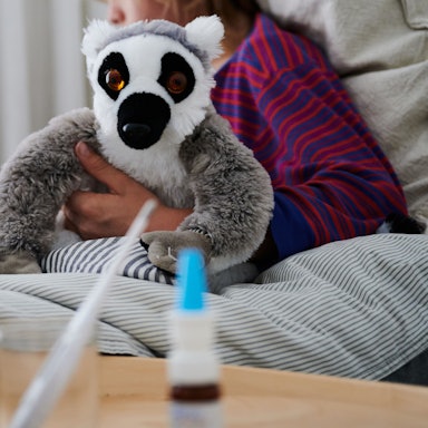 Eine Person liegt im Bett und guckt auf ihr Tablet, in der Hand ein Faultier-Kuscheltier. Auf dem Nachtisch stehen ein Fieberthermometer und ein Erkältungsspray.